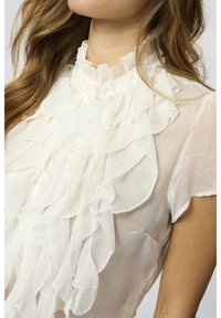 Blouse blanche à volants avec un col montant et une texture douce. Présente des volants superposés en cascade sur le devant et des manches courtes.