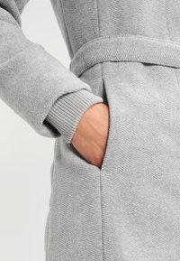 Abrigo de tejido texturizado gris con cinturón, cuenta con puños acanalados y un bolsillo lateral, mostrando una mano introducida en el bolsillo.