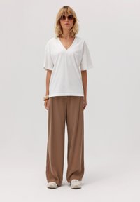Biała koszulka z dekoltem w serek, luźny krój, połączona z brązowymi spodniami o wysokim stanie i szerokich nogawkach. Modelka nosi beżowe skorupy i duże okulary przeciwsłoneczne.