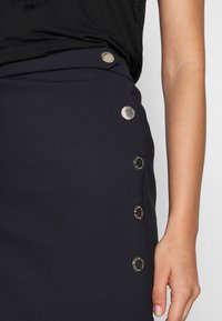 Närbild av en person som bär en svart kjol med fyra runda silverknappar längs sidan och en svart topp.