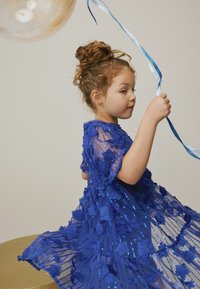Abito blu con paillettes, decorato con applicazioni floreali e strati di tessuto trasparente. Bambina che tiene dei nastri blu, con un palloncino sullo sfondo.