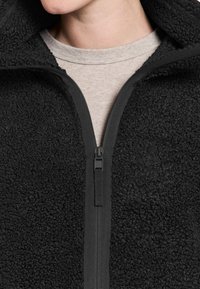 Sort fleecejakke med tekstureret overflade, lynlås og høj krave over en lys grå skjorte. Holdbart syntetisk materiale.