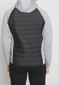 Bluza z kapturem w szarym kolorze z przodu i ciemnoszarym pikowanym tyłem z poziomymi liniami, wykonana z miękkiego materiału.