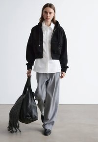 Schwarzer, gekürzter Hoodie mit silbernem Reißverschluss, weißes Hemd, graue Hosen mit weitem Bein und schwarze Tasche mit Fellakzenten. Glatte Texturen, lässiges Design.