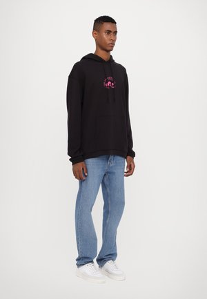 Zwarte hoodie met een roze logo, een voorzak en koordjes, gecombineerd met lichtblauwe jeans en witte sneakers. Effen achtergrond.
