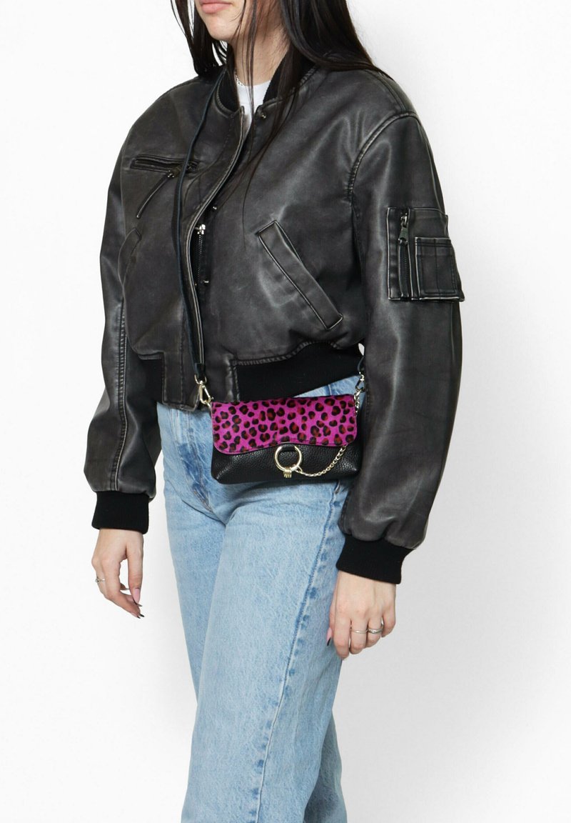 Giacca bomber nera in pelle con polsini a coste, abbinata a una piccola borsa a tracolla con stampa leopardata rosa e un accento in catena dorata. Jeans in denim chiaro.