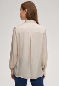 Lichtbeige blouse van zijde, met een hoge hals, lange mouwen, een plooi op de rug en een knoopdetail bij de hals. Gladde textuur.