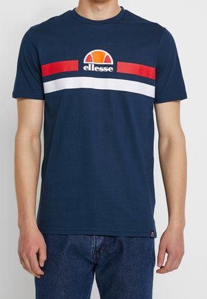 Homme portant un t-shirt ellesse bleu marine avec des rayures horizontales rouges et blanches sur la poitrine, associé à un jean bleu.