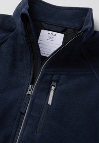 Navy fleece-takk med høy krage, frontglidelås og to glidelåslommer. Har en etikett med størrelse og vedlikeholdsinstruksjoner.