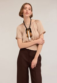 Blusa beige a maniche corte con scollatura arricciata, cintura testurizzata, abbinata a pantaloni larghi marrone scuro e una collana con nappine.
