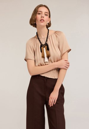 Beige short-sleeve blouse with a gathered neckline, textured waistband, paired with dark brown wide-leg trousers and a tassel necklace.