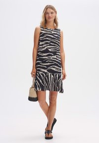 Ärmelloses Kleid in Schwarz mit weißen Zebra-Streifen; ausgestellter Saum; kombiniert mit einer geflochtenen Handtasche und schwarzen Sandalen; glatte Stoffstruktur.