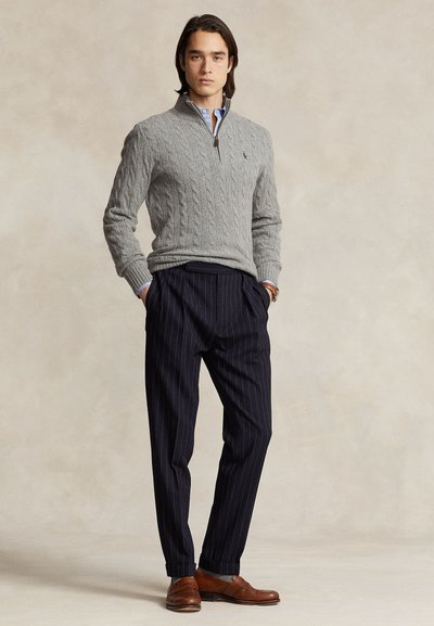 Uomo che indossa un maglione grigio a trecce, camicia azzurro chiaro, pantaloni blu navy a righe sottili e scarpe di pelle marroni, in piedi con le mani nelle tasche.