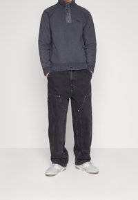 Sweat-shirt bleu marine zippé avec col montant, poignets côtelés, accompagné d'un pantalon noir ample avec poches avant et de baskets de couleur claire.