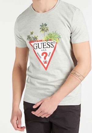 Man draagt een lichtgrijs GUESS T-shirt met een rood driehoeklogo, een vraagteken en palmbomen erboven, gecombineerd met een donkere broek.
