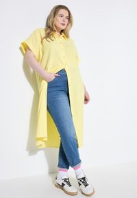 Camisa amarilla, de gran tamaño, abotonada y con mangas cortas encima de unos jeans azules. Zapatillas blancas con detalles negros y calcetas rosas y blancas visibles.