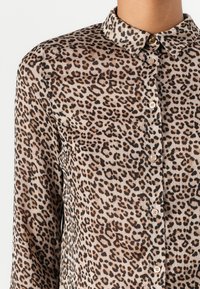 Leopardskinnet skjorte med krage, gjennomsiktig tekstur og knappelukking. Har varierte brune og svarte flekker på en lys beige bakgrunn.