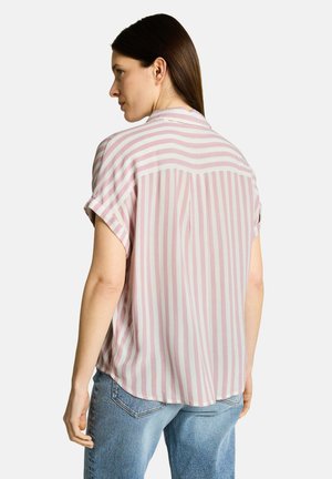 Femme faisant face à gauche, portant un chemisier à manches courtes rayé verticalement rose et blanc et un jean bleu clair, debout devant un fond uni.