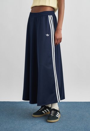 Kobieta w granatowej maxi spódnicy Adidas z białymi paskami i czarnych butach sportowych Adidas z białymi skarpetkami na niebieskim dywanie.