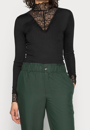 Haut noir à manches longues en côtelé avec des détails en dentelle sur la poitrine, le cou et les poignets, associé à un pantalon taille haute vert foncé, main dans la poche côté droit.