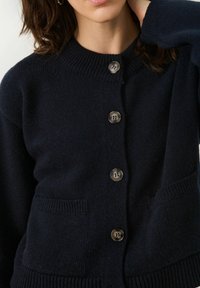 Navy knitted cardigan with a high collar, featuring five brown buttons and two front pockets. Soft texture and ribbed cuffs.