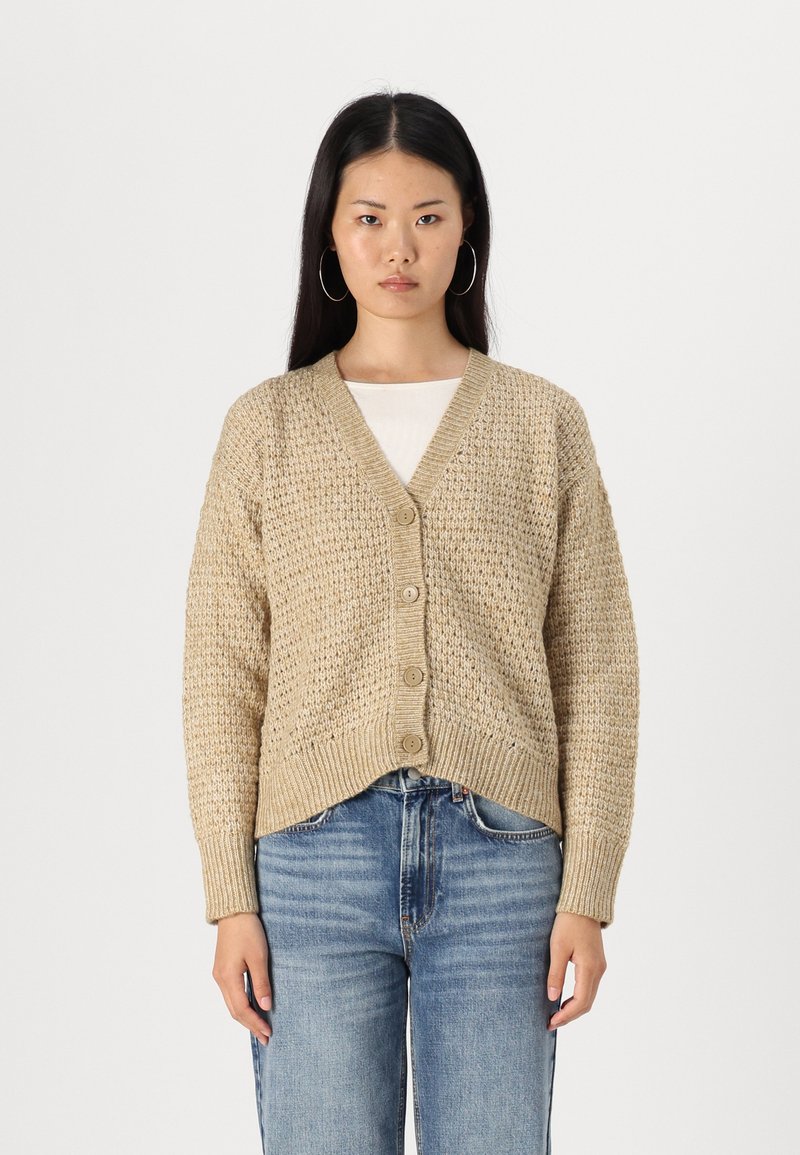 Beige strikket cardigan med V-hals, fem knapper og ribbede mansjetter, kombinert med blå jeans. Strukturert stoff, avslappet passform, uformell design.