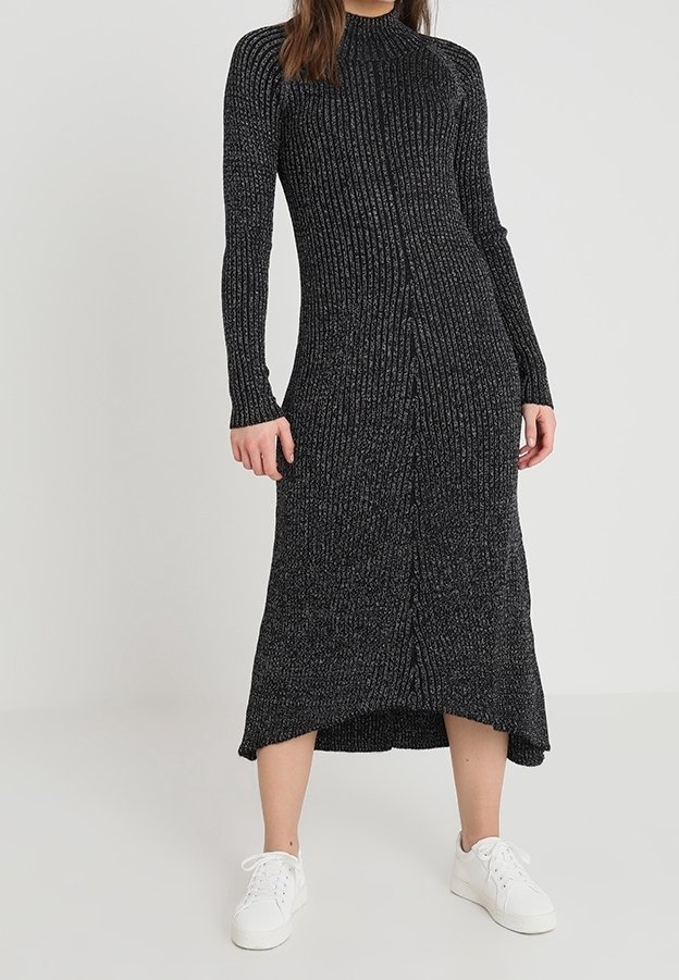 Femme portant une longue robe en maille côtelée noire à manches longues et des baskets blanches, debout devant un fond gris uni.