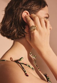 Zlati prstan v obliki školjke na roki, postavljen na kožo. Na rami so vidni trakovi s cvetličnim vzorcem. Mehka osvetlitev poudarja teksture.