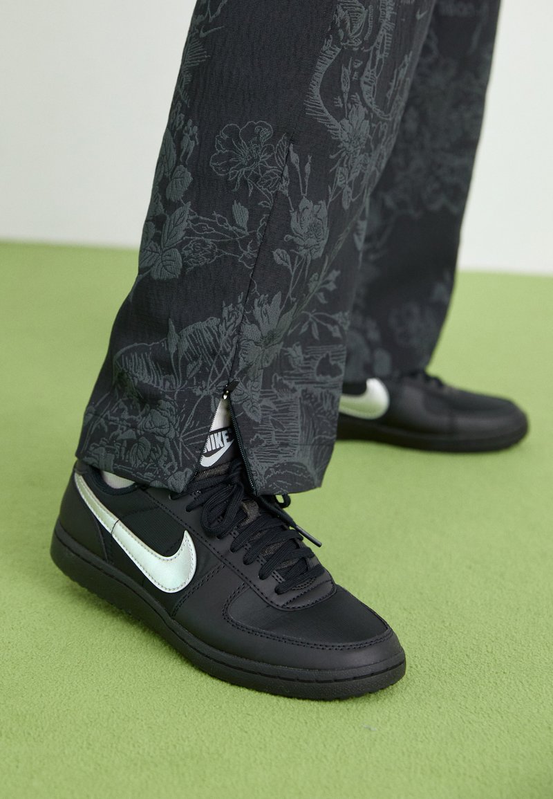 Sneaker nera con logo Nike argentato, caratterizzata da una tomaia in pelle liscia e suola in gomma testurizzata. Pantaloni con motivo floreale, in tessuto grigio scuro.