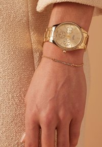 Gouden horloge met een metalen band en een ronde wijzerplaat, voorzien van drie subdialen en een met kristallen versierde bezel. Een delicate gouden armband is zichtbaar.