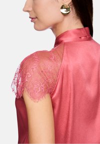 Satin-Oberteil in zartem Pink mit Spitzenkappenärmeln, ausgestattet mit einem hohen Kragen und glatter Textur, das feine Spitzenverzierungen an den Schultern hervorhebt.