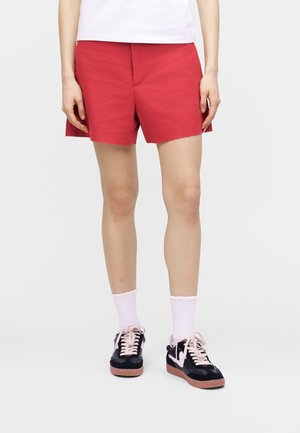 Henkilö, joka pukeutuu kirkkaanpunaisiin shortseihin, valkoisiin sukkiin ja mustiin lenkkareihin, joissa on vaaleanpunaiset nauhat, seisoo yksinkertaista vaaleaa taustaa vasten.