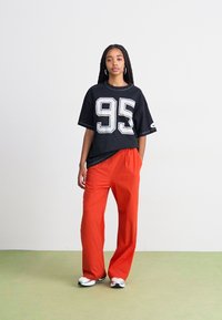 Svart oversized t-skjorte med hvit "95" trykk, parret med lys orange, løst sittende bukser og svart-hvite sneakers. Enkelt bakgrunn.