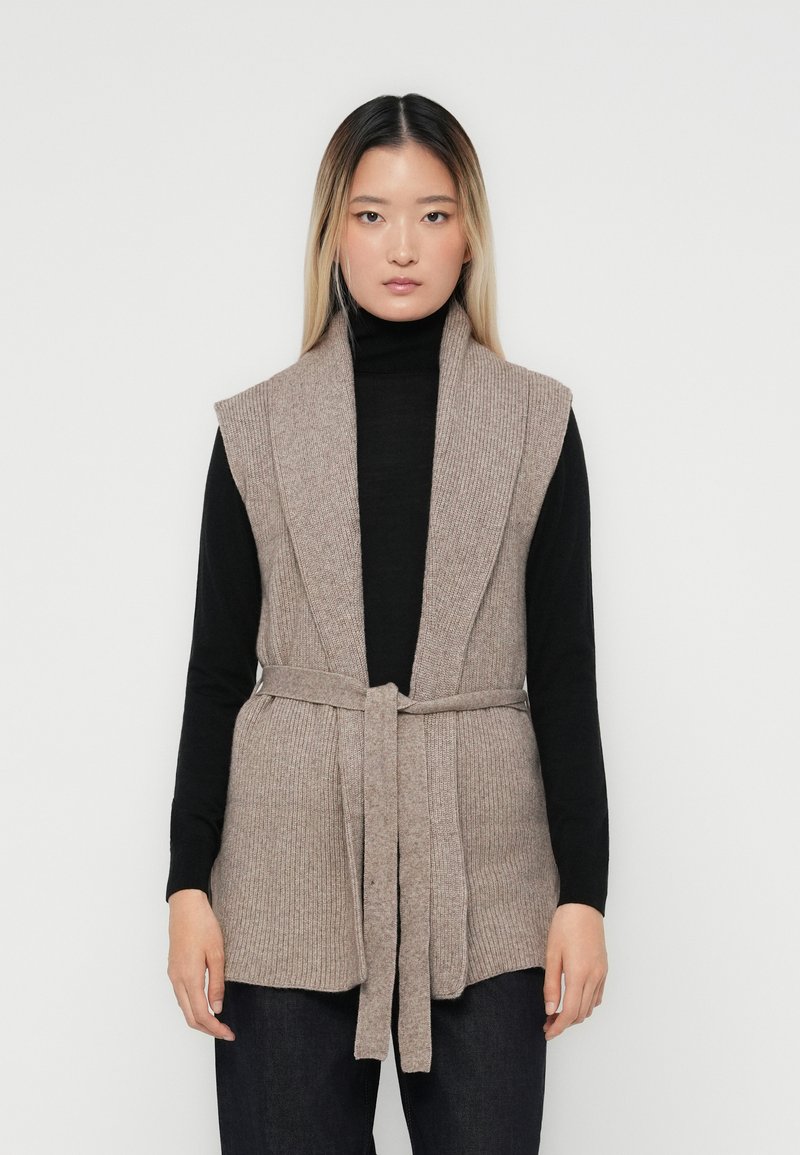 Beige geribde vest met brede revers en een riem in de taille, gedragen over een zwarte coltrui. De textuur is zacht, wat een gelaagd uiterlijk creëert.