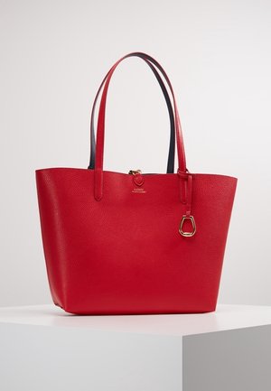 Rote Lederhandtasche mit Textur, zwei langen Henkeln, goldfarbenen Metallakzenten und einem kleinen hängenden Anhänger, präsentiert auf einem weißen Podest.