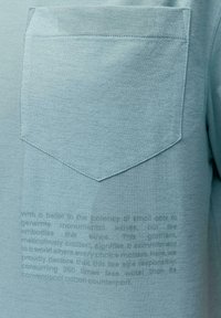 Šviesiai mėlyna marškinėliai su tekstūruotu audiniu, su maža kišenėle ir iškiliais tekstiniais elementais apatinėje priekyje.