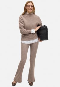 Suéter marrón de punto acanalado con cuello alto, combinado con pantalones de campana a juego. Camisa blanca debajo, sosteniendo un bolso negro. Zapatos de estampado animal.