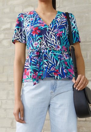 Femme portant un chemisier cache-cœur bleu à motifs floraux avec des manches courtes et un jean bleu clair, tenant un sac à épaule noir sur un fond de brique.