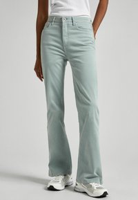 Light green flared jeans with a button and zip fly closure, front pockets, and a smooth, slightly textured fabric, paired with white athletic sneakers.