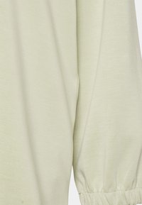 Ljusgrön, mjuk tyg med en slät textur. Har långa ärmar med elastiska manschetter. Enkelt design, inga mönster eller utsmyckningar.