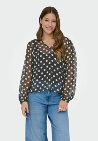 Jeune femme souriante, portant un blouse noire transparente à pois noirs par-dessus un top noir et un jean large bleu clair, se tenant devant un fond blanc.
