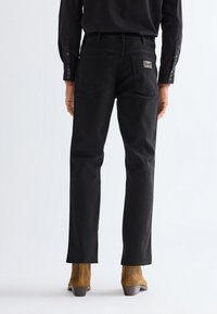 Person wearing black straight-leg jeans with visible back pockets and tan suede ankle boots, standing on a plain white background.