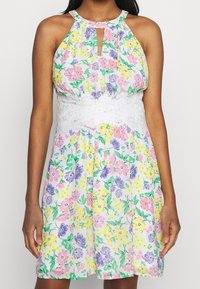 Robe fleurie avec une ceinture en dentelle blanche, présentant un col licou et une découpe en forme de goutte. Les couleurs incluent le rose, le jaune et le violet.