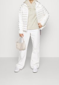 Chaqueta acolchada blanca, sudadera gris claro con logotipo, pantalones anchos blancos y zapatillas blancas. Sostiene un pequeño bolso de mano texturizado en beige.