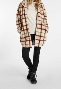 Pluche beige jas met een rood en zwart geruite patroon, oversized ontwerp en capuchon; gecombineerd met een crèmekleurig ribbeltop en zwarte broek.