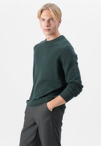 Pulover tricotat de culoare verde închis, cu guler rotund și manșete cu nervuri, asortat cu pantaloni gri îmbrăcați pe corp. Textură netedă, croială casual.