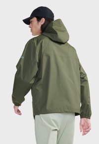 Olijfgroene waterdichte jas met een capuchon, elastische manchetten en een eenvoudig ontwerp. Achteraanzicht met een zwarte pet en lichtgekleurde broek.