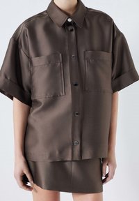 Brązowa satynowa koszula z krótkim rękawem z kołnierzykiem, z dwiema przednimi kieszeniami i czarnymi guzikami. Gładka tekstura i luźny krój, zestawiona ze spódnicą w tym samym kolorze.