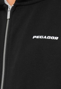 Felpa nera con zip realizzata in tessuto morbido, con zip argentata e logo bianco "PEGADOR" sulla parte sinistra del petto.