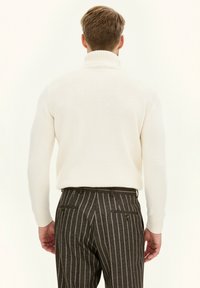 Kremowy sweter z golfem z prążkowanym kołnierzem, zestawiony z ciemnymi spodniami w paski z kieszeniami i guzikami z tyłu.